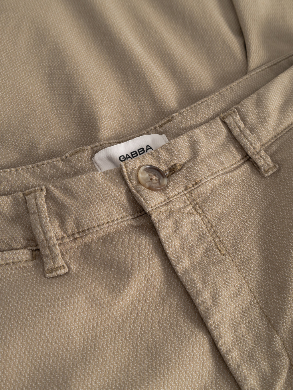 Close-up of GABpaul K3280 Dale Chino in sand by Gabba, showing beige textured fabric, button closure, belt loops, French pockets, and a partially unbuttoned waistband revealing a white Gabba label inside.