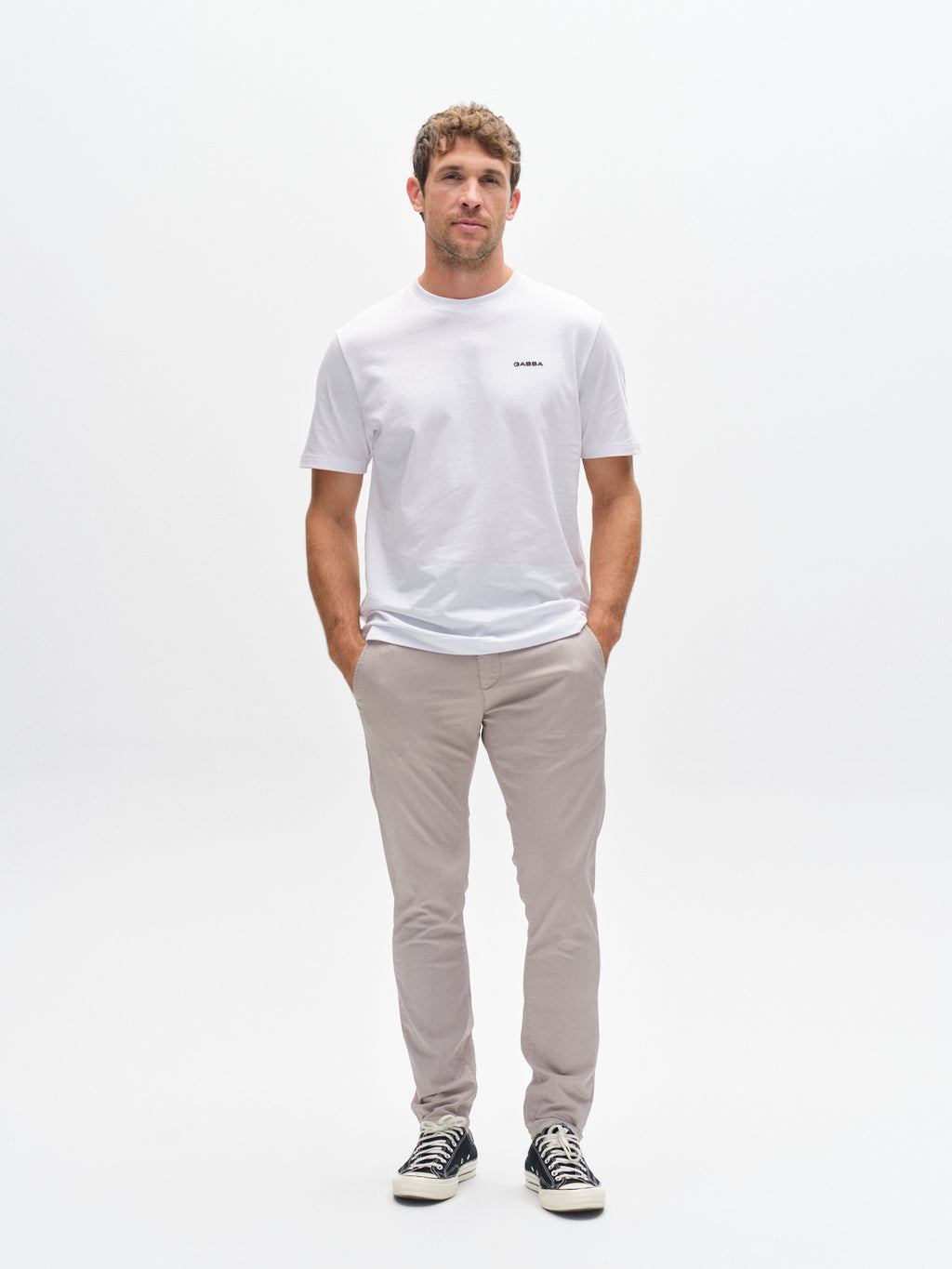 A man stands against a plain white background wearing a white t-shirt, black sneakers, and Gabba GABpaul K3280 Dale Chino in Grey Paloma with French pockets, hands in his pockets and looking straight ahead neutrally.