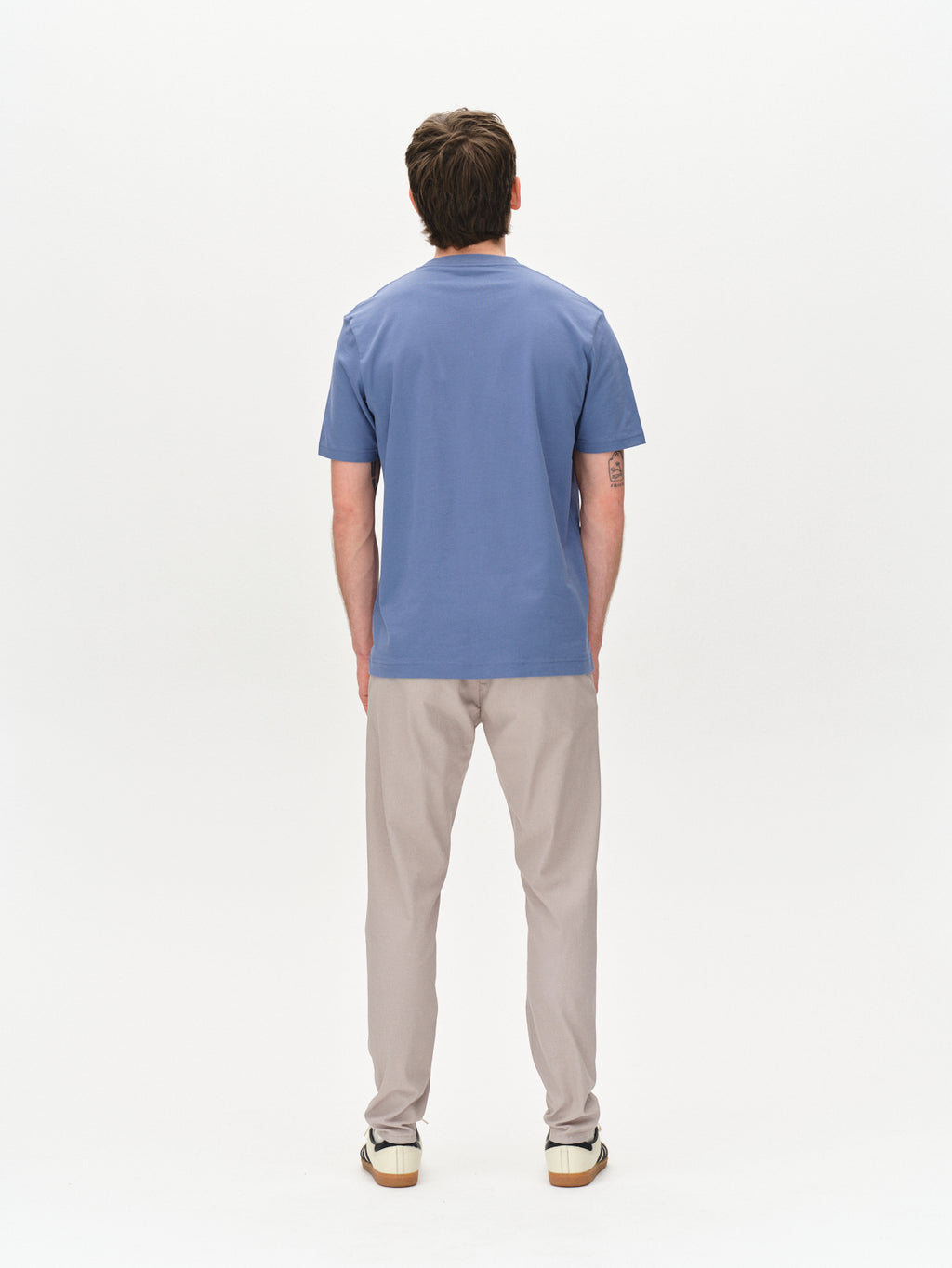 A person stands facing away from the camera, wearing a short-sleeve blue shirt, white sneakers, and Gabba Paul K3280 Dale Chino in Grey Paloma, against a plain white background.