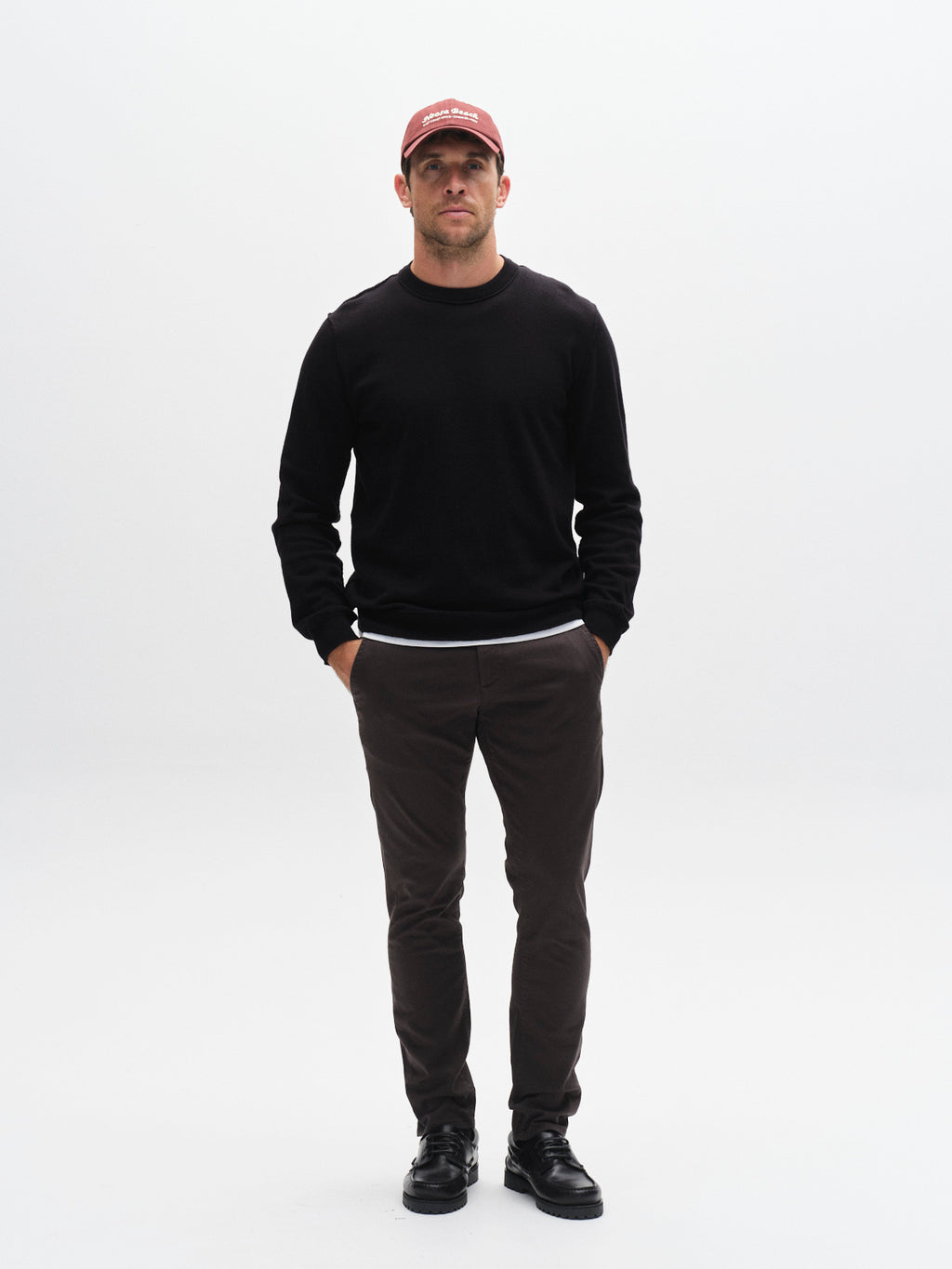 A man stands facing the camera against a white background. He wears a red cap, black long-sleeve shirt, Gabba GABpaul K3280 Dale Chino - Charcoal pants with French and double paspel back pockets, and black shoes, hands in his pockets.