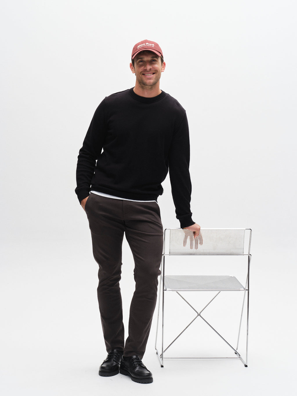 A man in a red cap, black sweater, Gabba GABpaul K3280 Dale Chino pants in charcoal, and black shoes smiles with one hand on a metal chair against a plain white background.