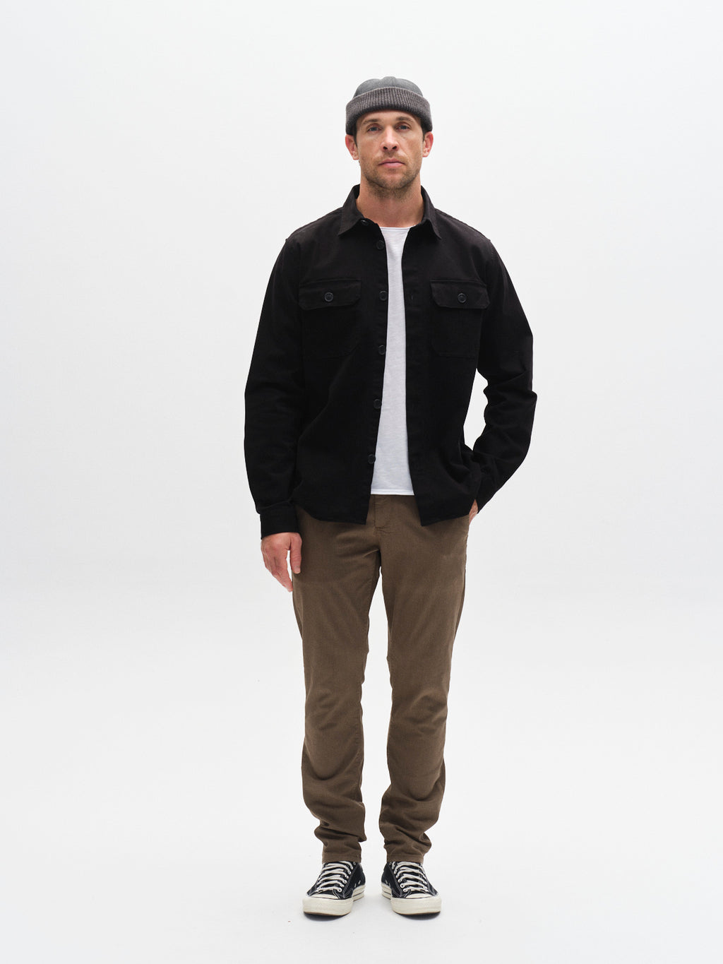 A man wears a gray beanie, black button-up shirt, white t-shirt, Gabba GABpaul K3280 Dale Chino - Crocodile slim pants with French pockets, and black-and-white sneakers while standing against a plain white background.