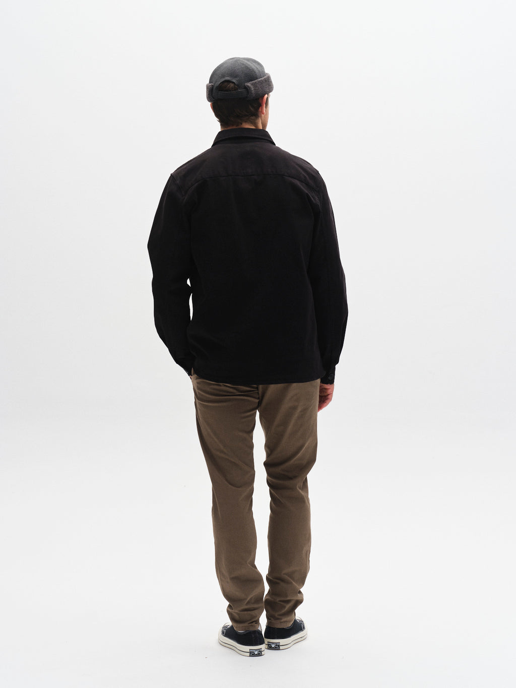 A person in a gray cap, black jacket, Gabba GABpaul K3280 Dale Chino - Crocodile pants with French pockets, and sneakers stands facing away on a plain white background.