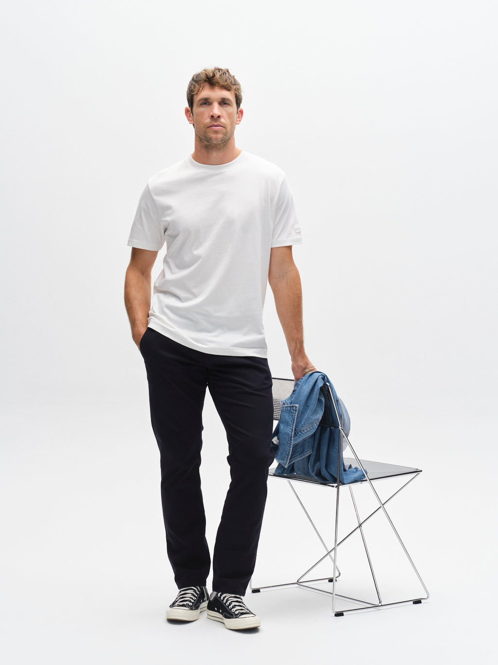 A man in a white t-shirt, Gabba GABpaul K3280 Dale Chino - Blue Black pants, and black sneakers stands against a plain white background with one hand in his pocket and the other on a chair holding a folded blue denim jacket.