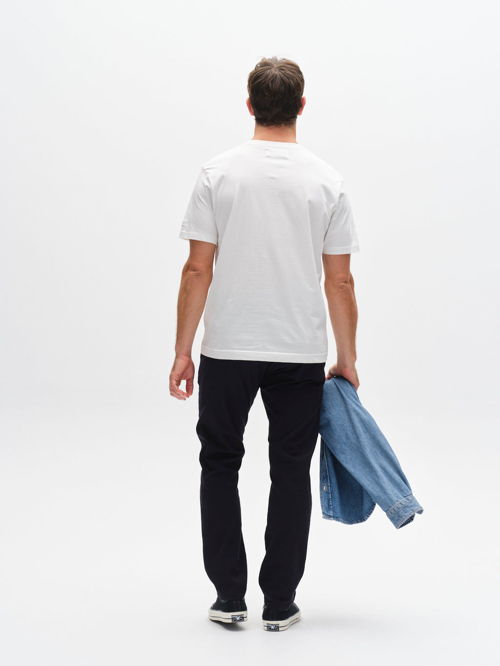 A man stands with his back to the camera, wearing a white t-shirt, Gabba GABpaul K3280 Dale Chino - Blue Black pants with double paspel back pockets, sneakers, and holding a denim jacket in his left hand against a plain white background.