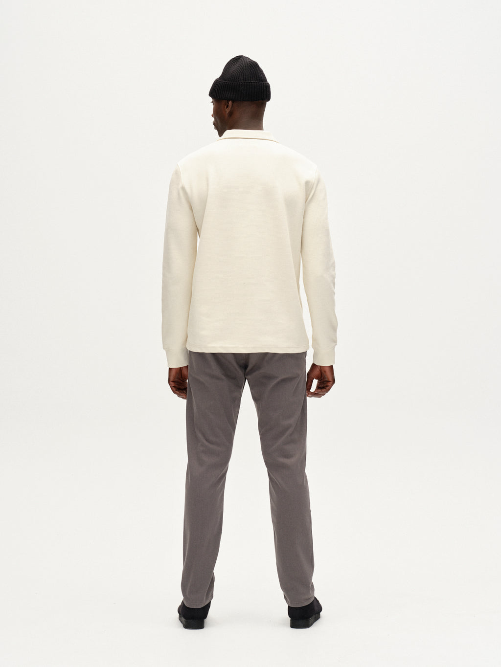 A man stands facing away, wearing a black beanie, a cream long-sleeve shirt, Gabba Paul K3280 Dale Chino pants in Dust Sage Green with French pockets, and black shoes against a plain white background.