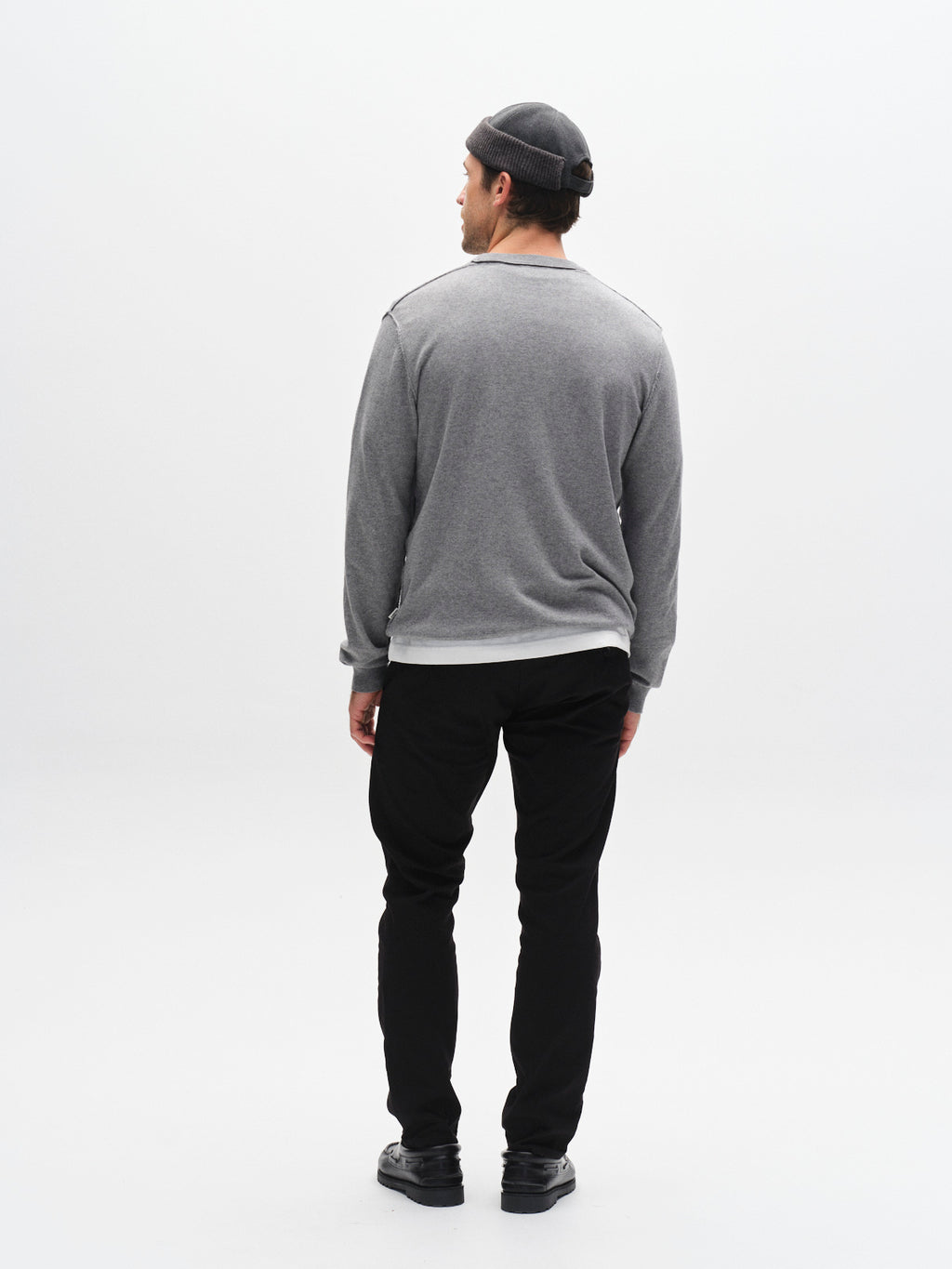 A man stands facing away from the camera, wearing a grey long-sleeve shirt, GABBA GABpaul K3280 Dale Chino - Black with French pockets and double paspel back pockets, black shoes, and a grey beanie against a plain white background.