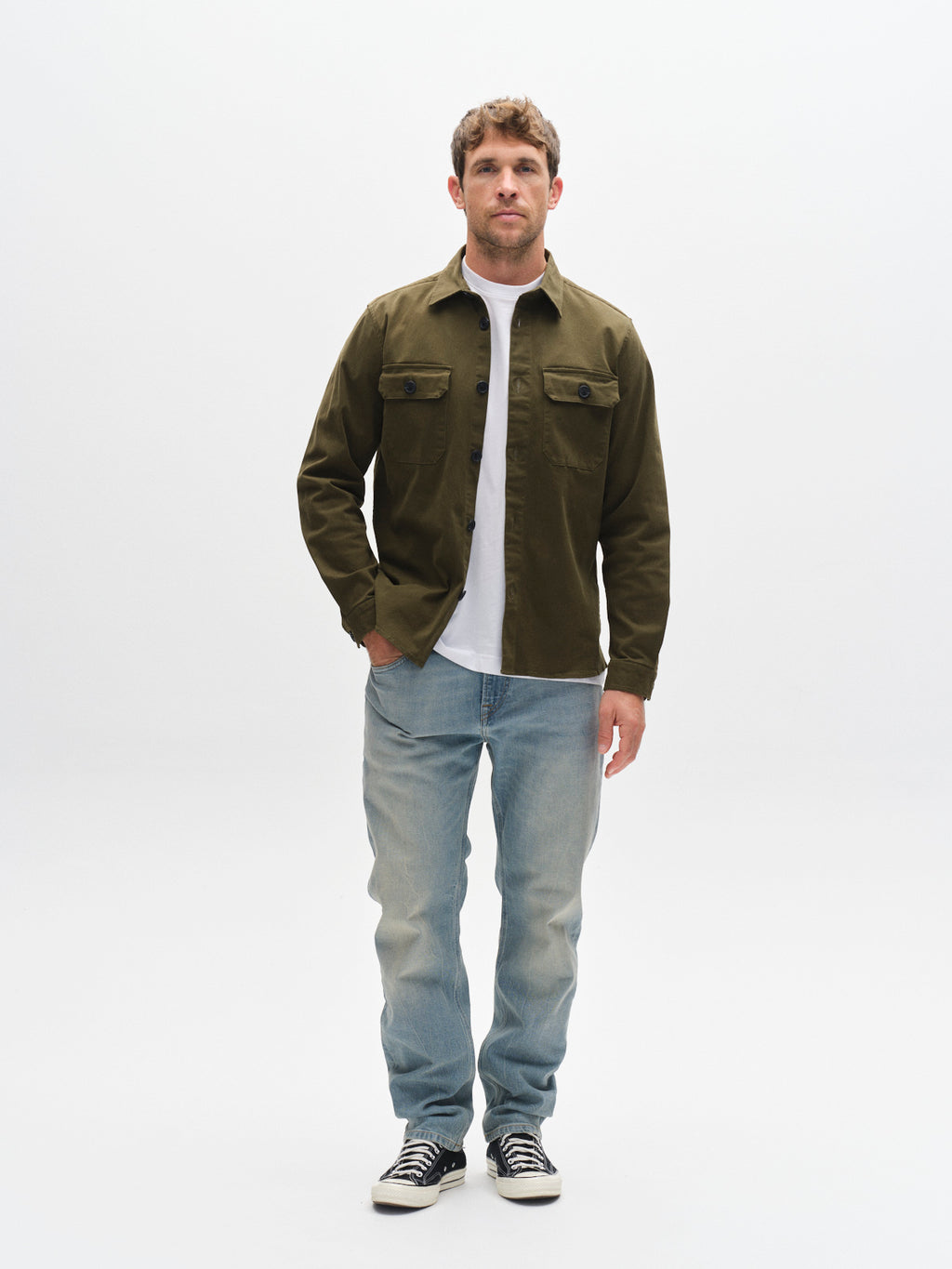 A man stands facing forward against a plain white background, wearing the GABBA Topper LS Shirt in Army with chest pockets over a white t-shirt, paired with light blue jeans and black-and-white sneakers.