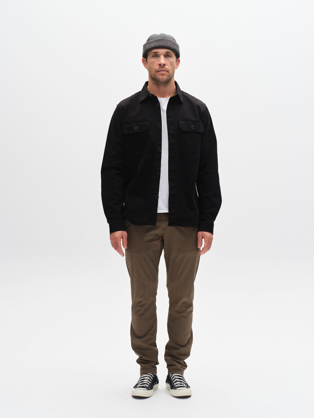 A man faces forward against a white background, wearing the GABBA Topper LS Shirt in black with chest pockets over a white collared shirt, paired with a gray beanie, brown pants, and black-and-white sneakers.