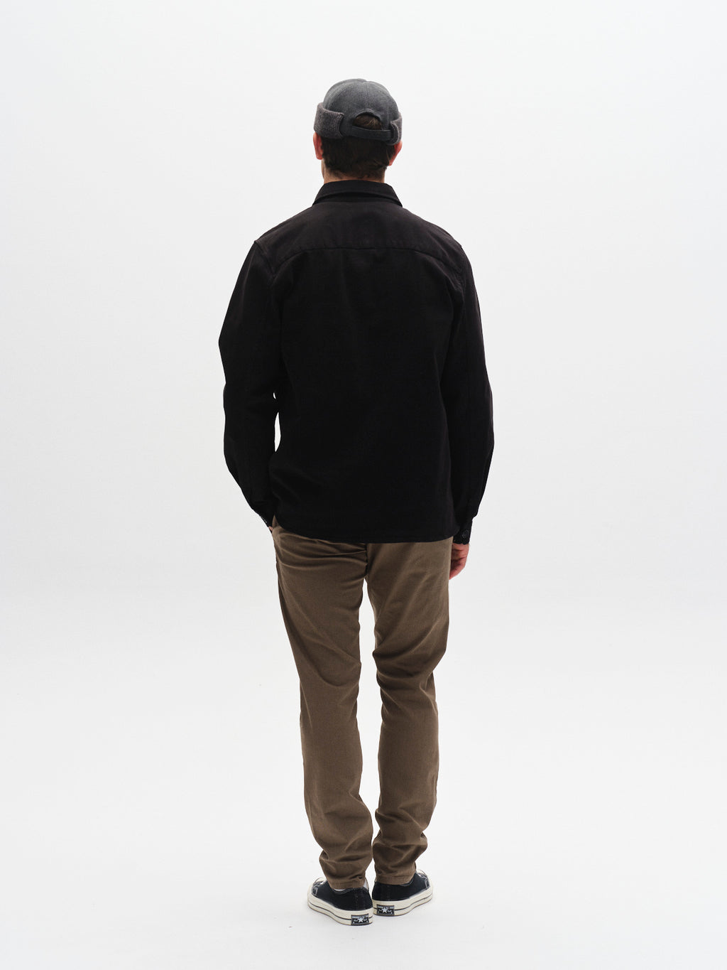 Wearing the GABBA Topper LS Shirt in Black styled as an overshirt, a person faces away from the camera against a plain white background, effortlessly layering with brown pants and sneakers.