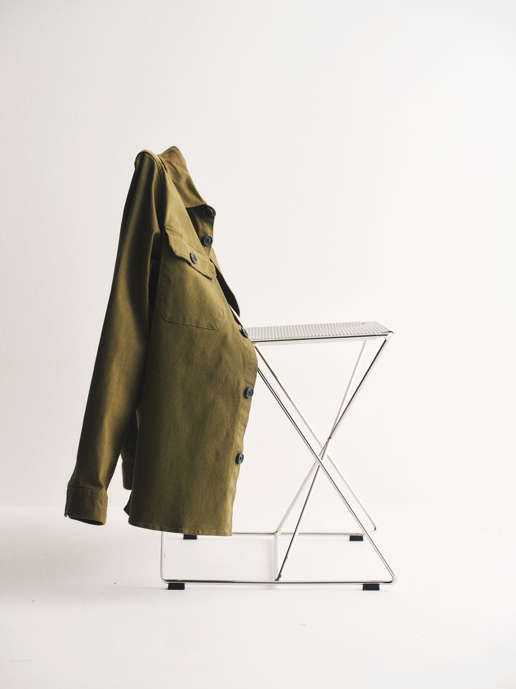 The GABBA Topper LS Shirt - Army, an olive green overshirt with chest pockets, is draped over a minimalist metal folding chair against a plain white background.