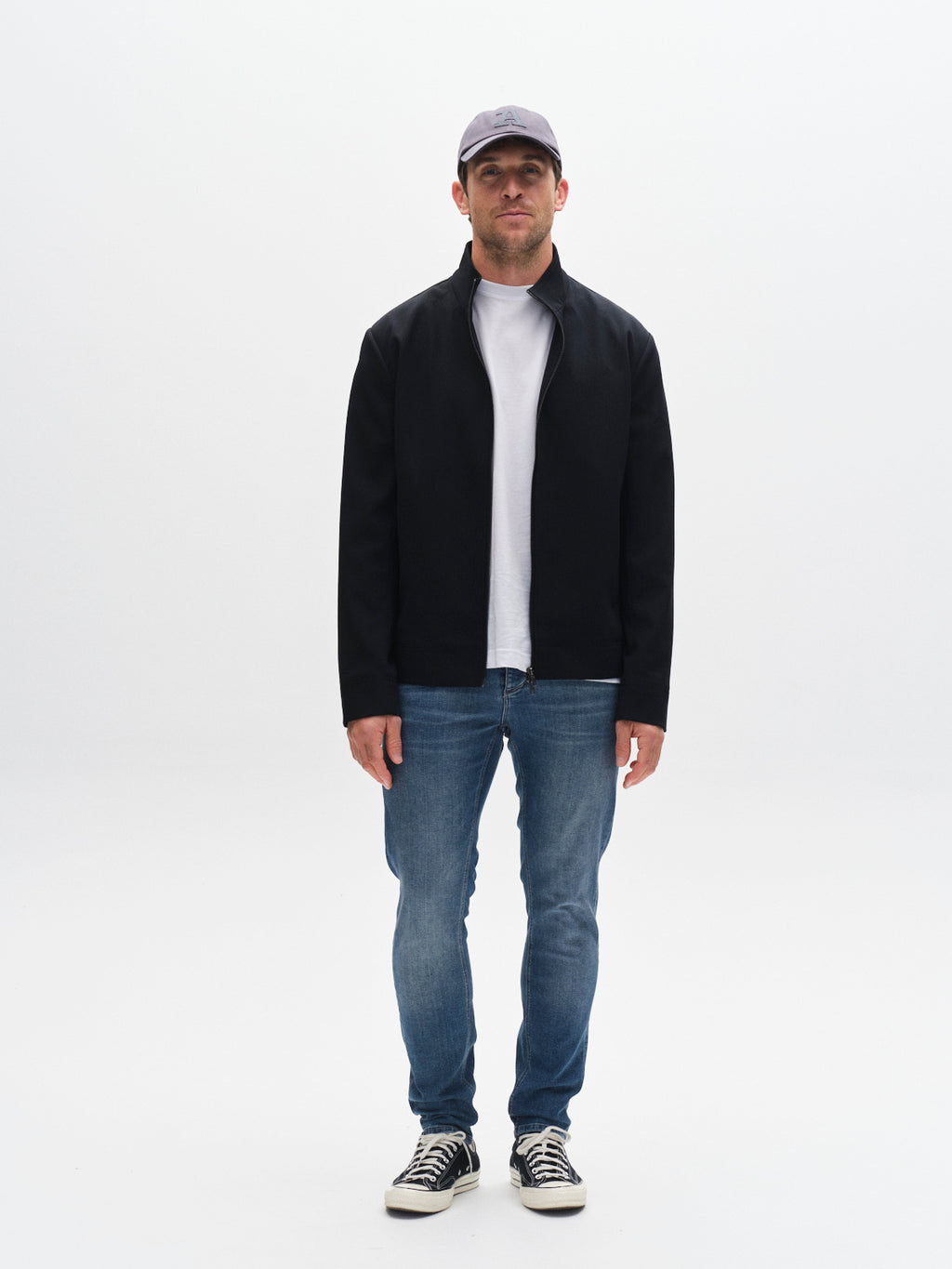A man stands facing forward against a plain white background, wearing a gray baseball cap, black jacket, white t-shirt, GABBA GABrey K3866 TENCEL™ Jeans in denim wash, and black-and-white sneakers for a laid-back look.