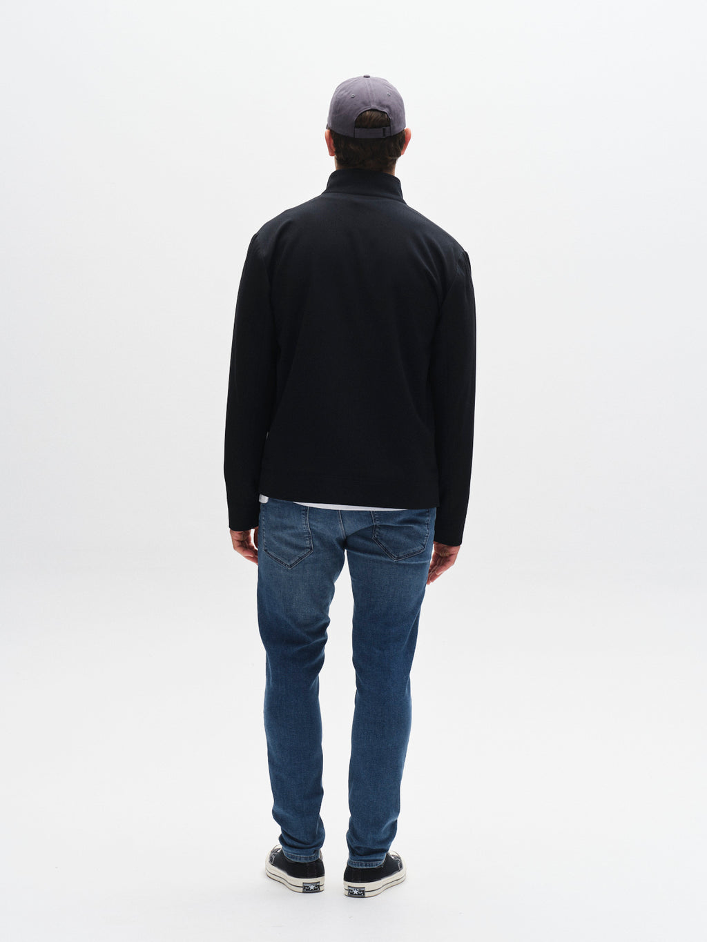 A man faces away from the camera in a black jacket, GABBA GABrey K3866 TENCEL™ Jeans in denim wash, black and white sneakers, and a gray cap, presenting a relaxed style against a plain white backdrop.