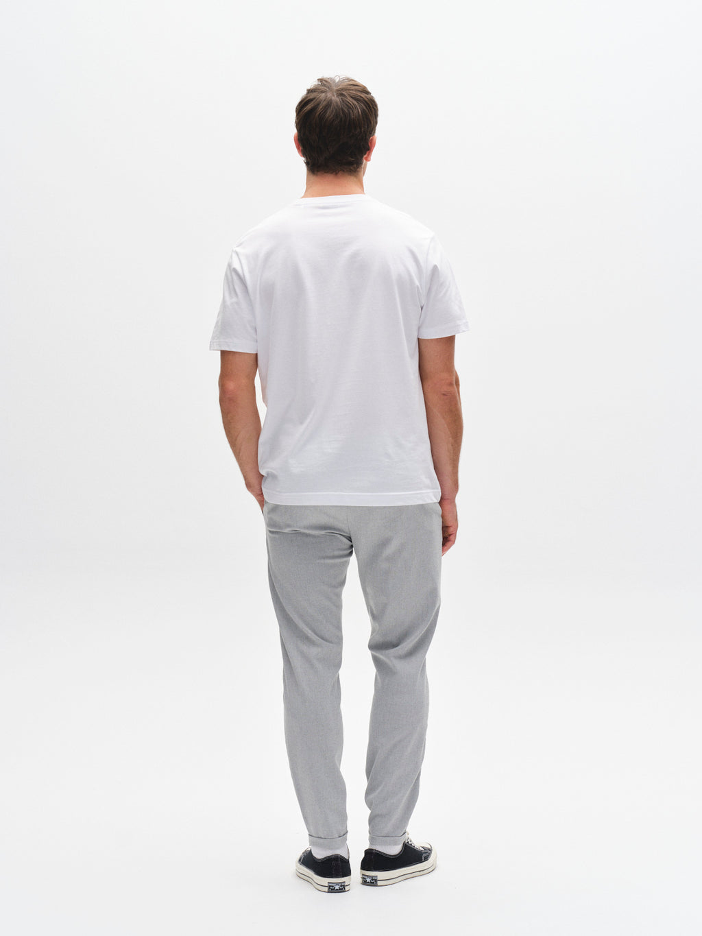 A man stands facing away from the camera against a white background, wearing a plain white t-shirt, Gabba Pisa Domo Pants in Monument, and black slip-on shoes.
