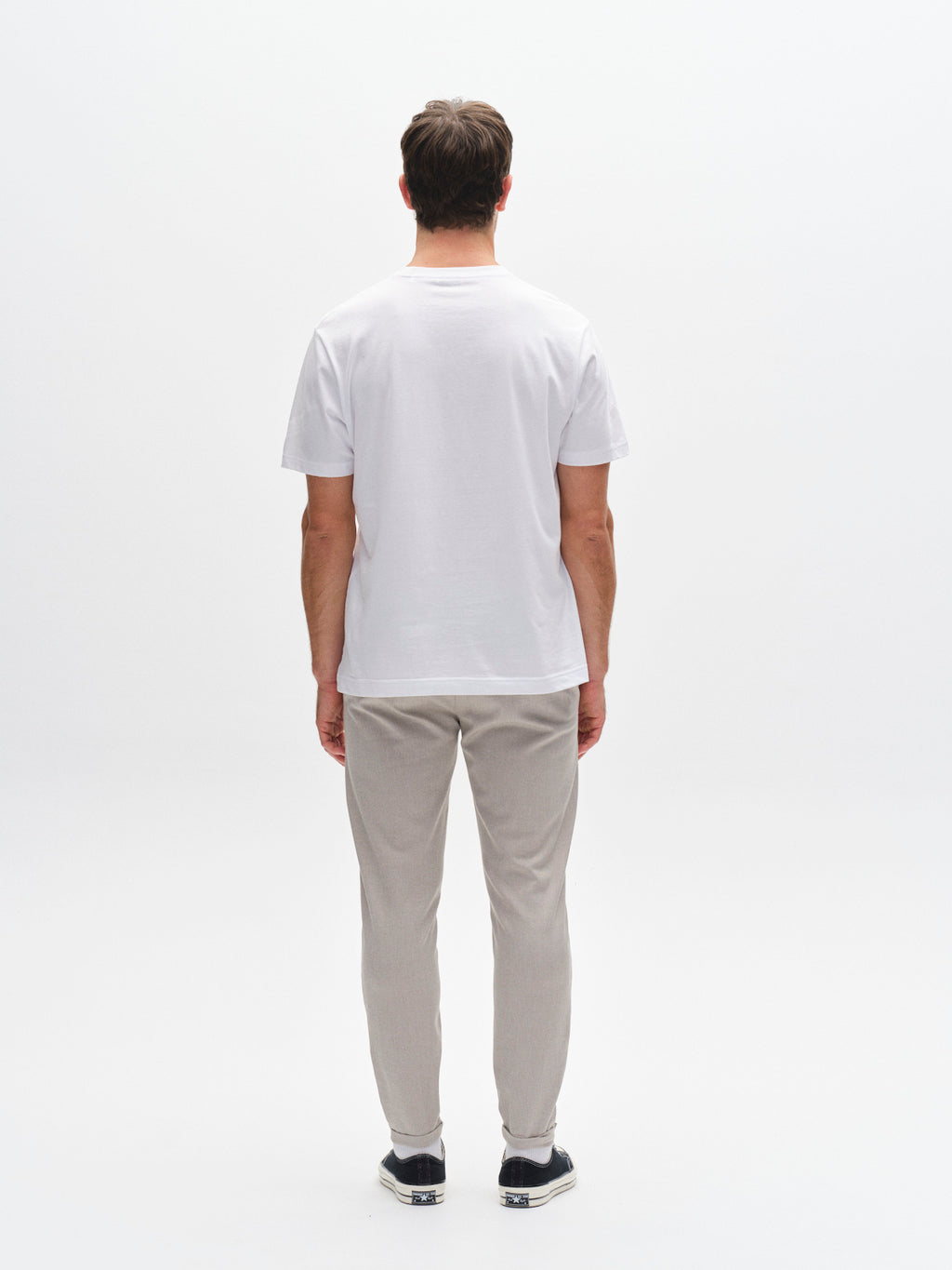 A man faces away from the camera against a white background, wearing a white t-shirt, Gabba Pisa Domo Pants in Coriander, and black slip-on shoes with white soles for a relaxed metro style.