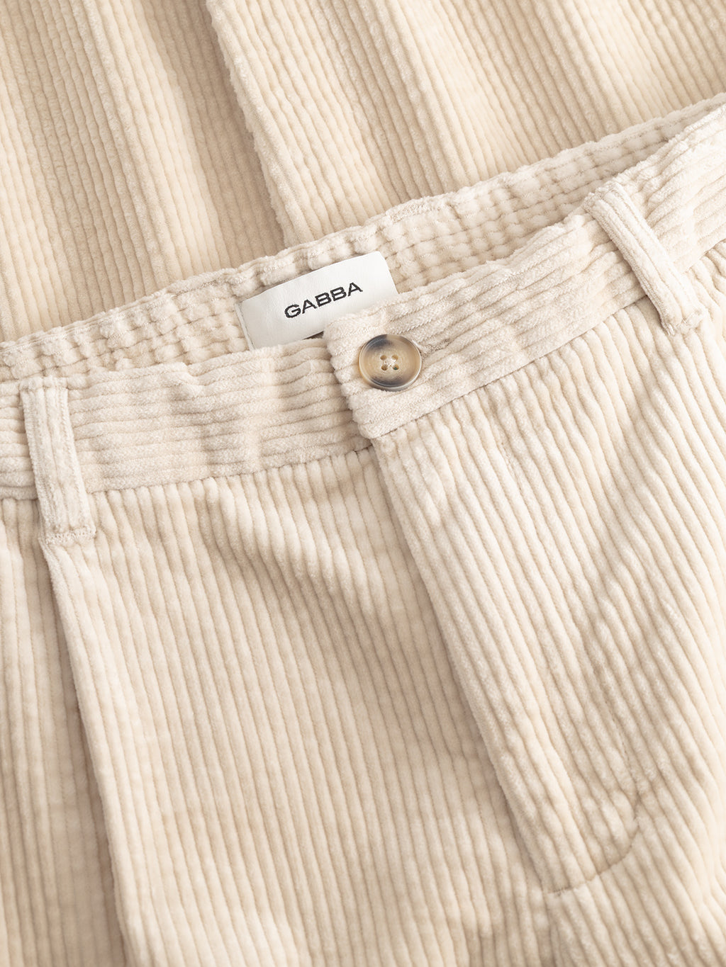 Close-up of the GABnapoli 19 Ibre Pant in Birch by Gabba, featuring beige cotton corduroy fabric, a button closure, belt loops, and a white label displaying the Gabba brand name. These men’s tailored pants offer a classic straight fit for versatile styling.