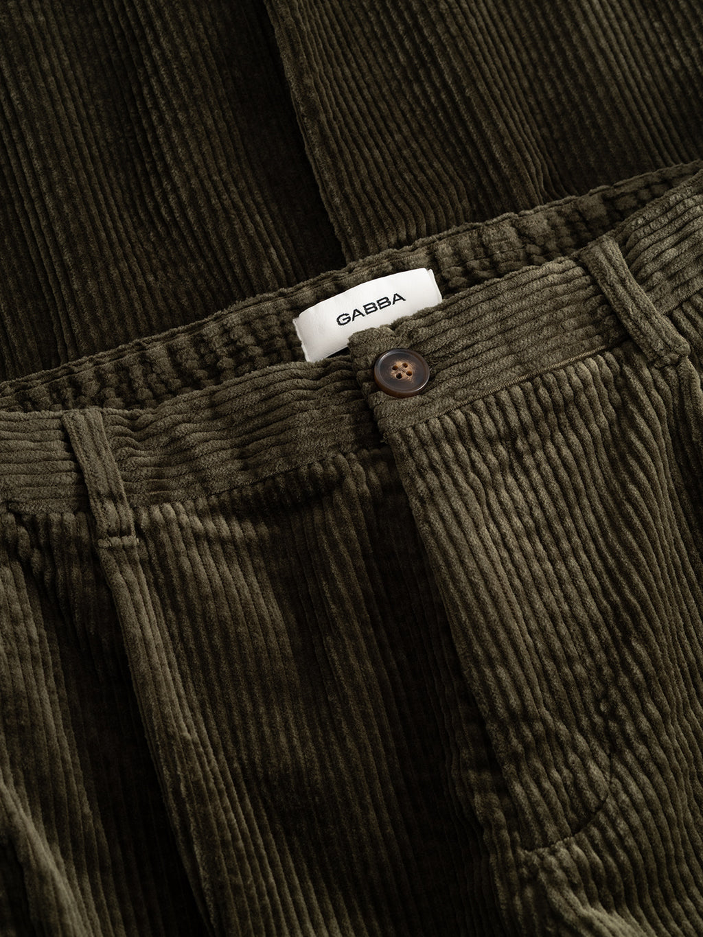 Close-up of the GABnapoli 19 Ibre Pant in Forest Night by Gabba, showcasing olive green cotton corduroy fabric with a brown button, elastic waistband, and a white Gabba label visible inside. The straight fit creates a tailored silhouette for effortless style.