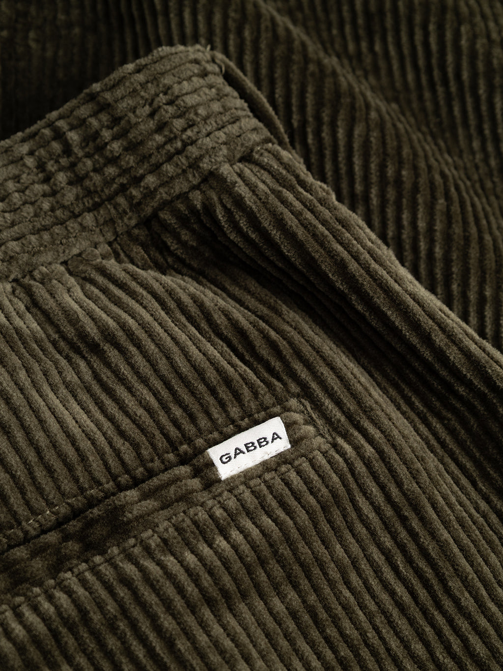 Close-up of the GABnapoli 19 Ibre Pant in Forest Night by Gabba, showcasing dark olive green cotton corduroy fabric with a visible pocket and a small white label that reads “Gabba.” The ridged texture highlights its straight fit and tailored silhouette.