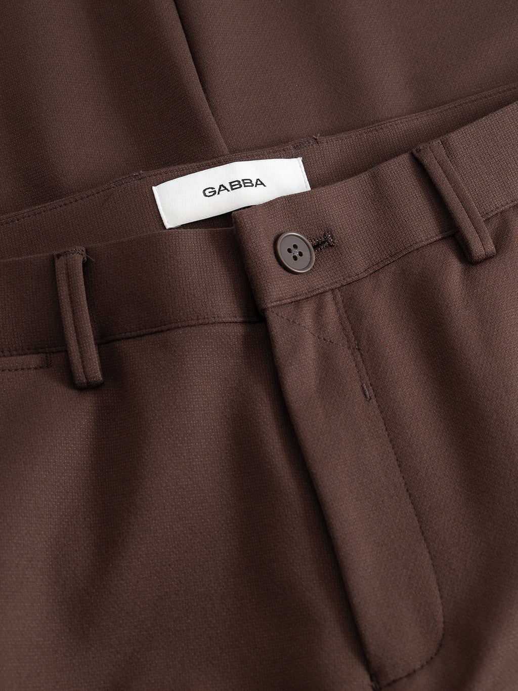 Close-up of the GABpaul Dotty Pant in Brown Slate by Gabba, featuring a button and zipper closure. The inner label displays the Gabba branding. Designed in a slim chino fit, the tailored fabric appears smooth, with belt loops along the waistband for added functionality.
