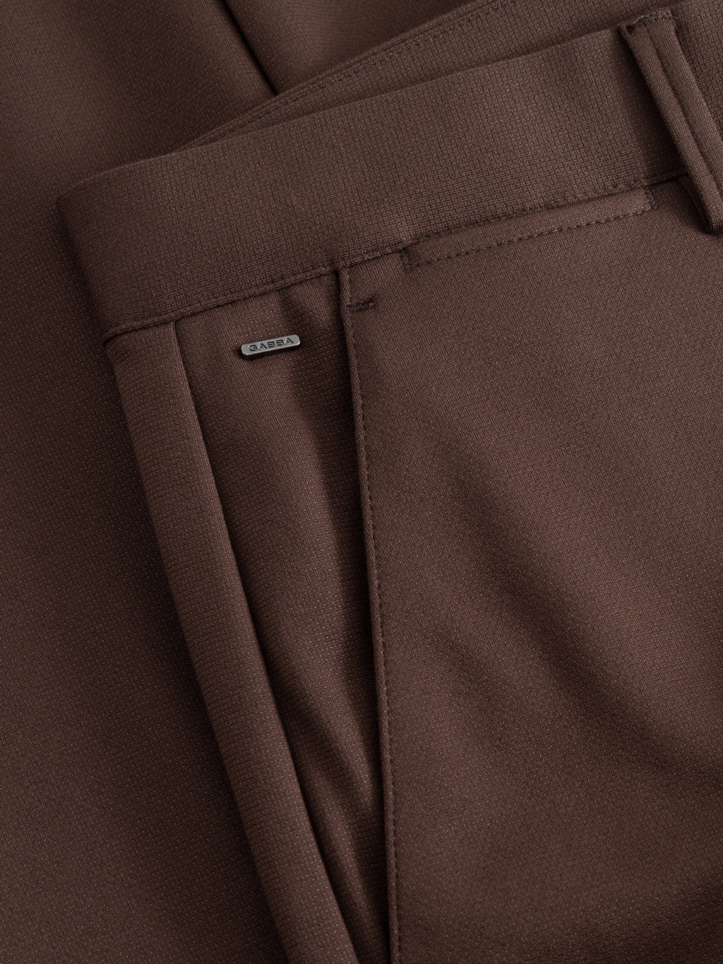 Close-up of the GABpaul Dotty Pant in Brown Slate, highlighting the pocket area. A small metallic tag with the brand name Gabba is attached near the pocket opening. The slim chino fit accentuates the smooth and slightly textured fabric, which features a subtle micro-pattern weave for added sophistication.