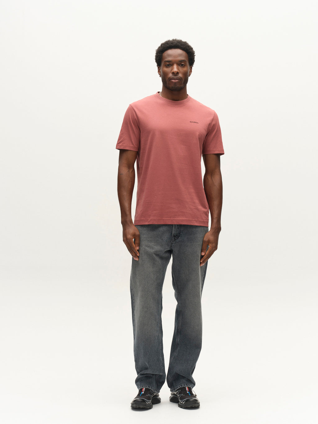 A man stands facing forward, wearing the Gabba GABdune Logo GOTS SS T-Shirt in Brick Red, paired with faded blue jeans and black sneakers against a plain, light background.