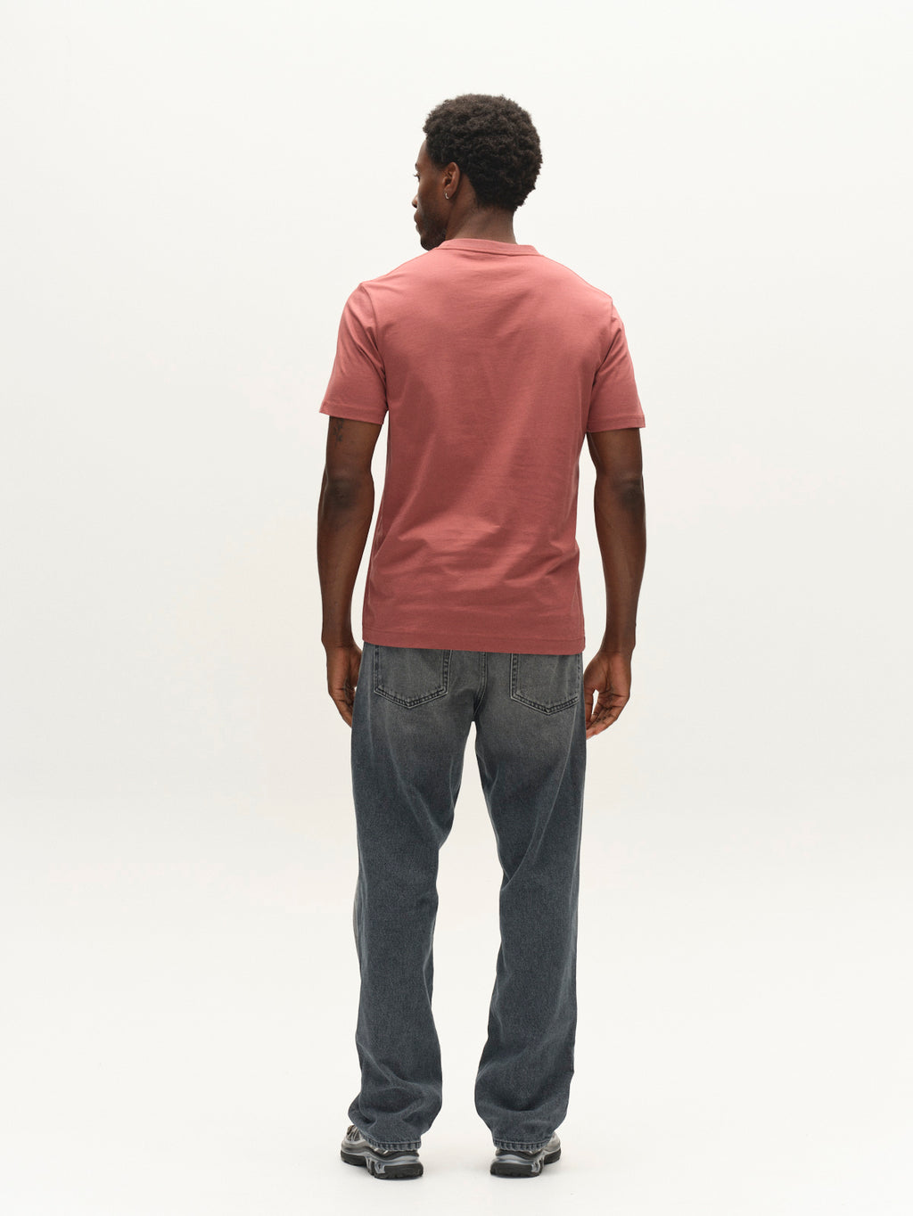 A man stands facing away, wearing the Gabba GABdune Logo GOTS SS T-Shirt in Brick Red, paired with blue jeans and sneakers, against a plain white background.