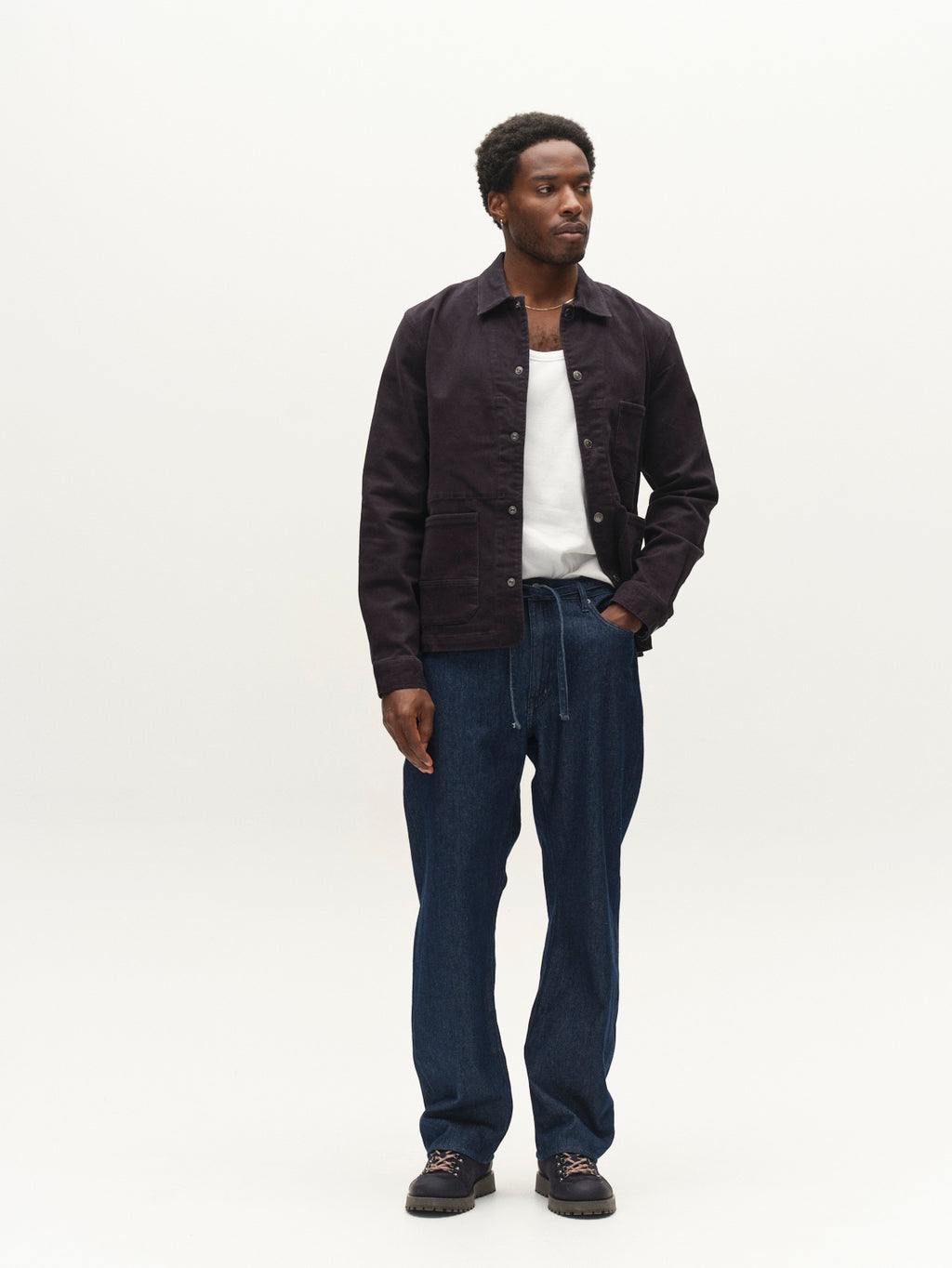 A man stands against a plain white background, wearing the Gabba GABbeta Twill - Navy jacket over a white shirt, regular fit blue jeans with a drawstring and utility pockets, and dark shoes. He looks slightly to the side with his hands relaxed by his sides.