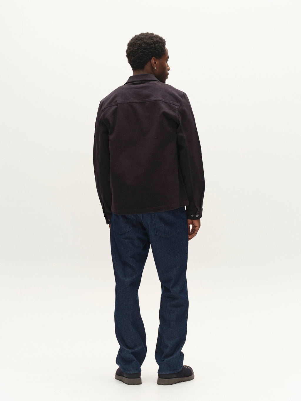 A man stands facing away from the camera, wearing a dark jacket and Gabba GABbeta Twill - Navy jeans crafted from garment-dyed stretch twill with a regular fit, paired with black shoes against a plain, light background.