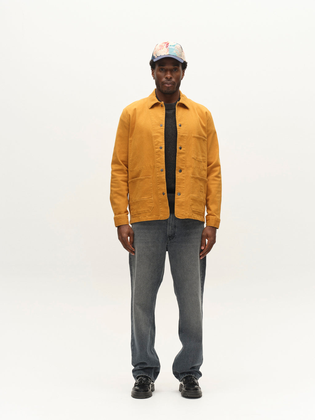 A man wearing the Gabba GABbeta Twill jacket in mustard yellow, featuring utility pockets, paired with a dark shirt, regular-fit gray jeans, black shoes, and a colorful cap stands facing the camera against a plain white background.