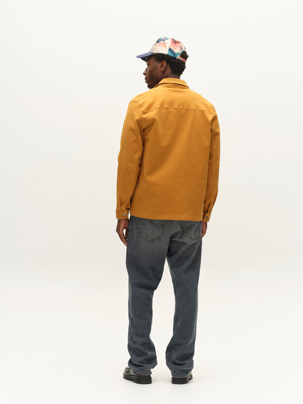 A person stands facing away, wearing the Gabba GABbeta Twill jacket in mustard yellow with utility pockets, paired with loose blue jeans, dark shoes, and a colorful cap against a plain white background.