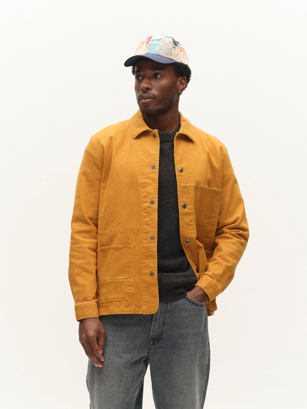A man stands against a plain white background, looking to the side, dressed in the Gabba GABbeta Twill - Mustard Yellow jacket with utility pockets, a dark sweater, regular fit jeans, and a multicolored cap.