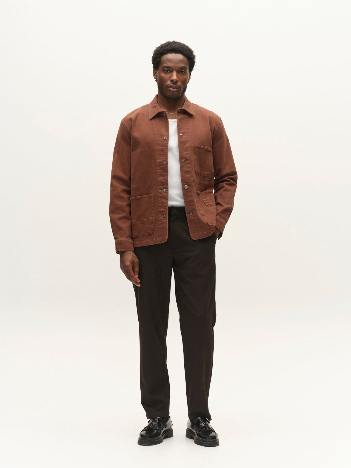 A man stands facing forward, wearing the Gabba GABbeta Twill jacket in Brown Slate with utility pockets over a white shirt, paired with dark trousers and black shoes, against a plain light background.