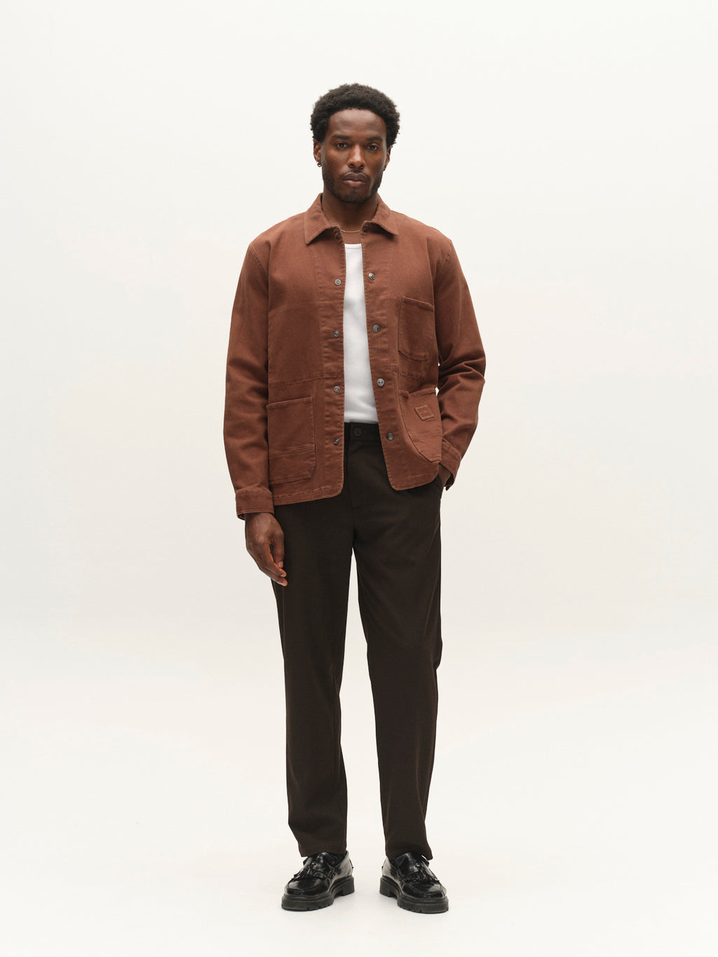 A man stands facing forward, wearing the Gabba GABbeta Twill jacket in Brown Slate with utility pockets over a white shirt, paired with dark trousers and black shoes, against a plain light background.