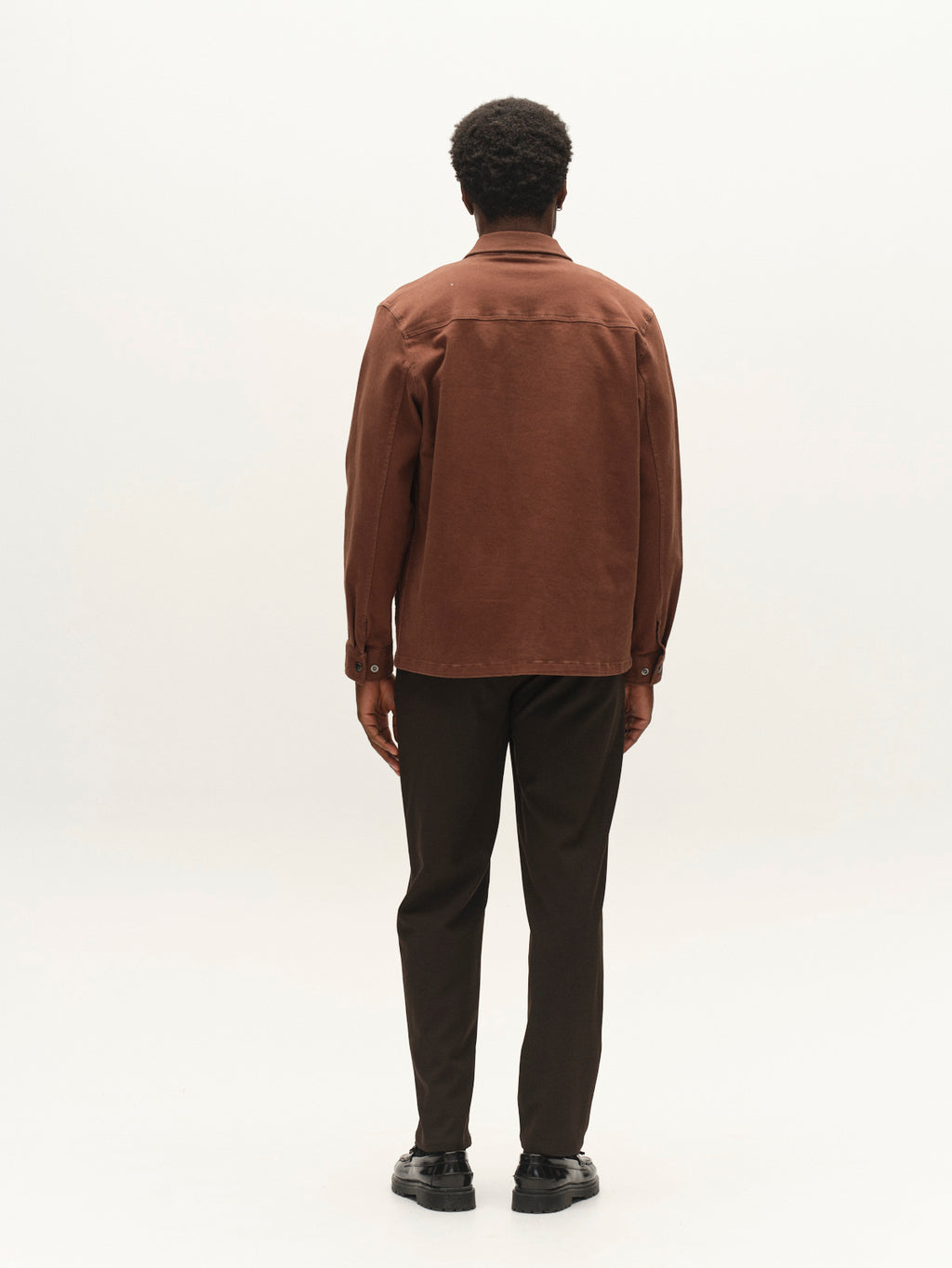 A person stands facing away from the camera, wearing the Gabba GABbeta Twill - Brown Slate jacket with utility pockets, paired with dark pants and black shoes, against a plain white background.
