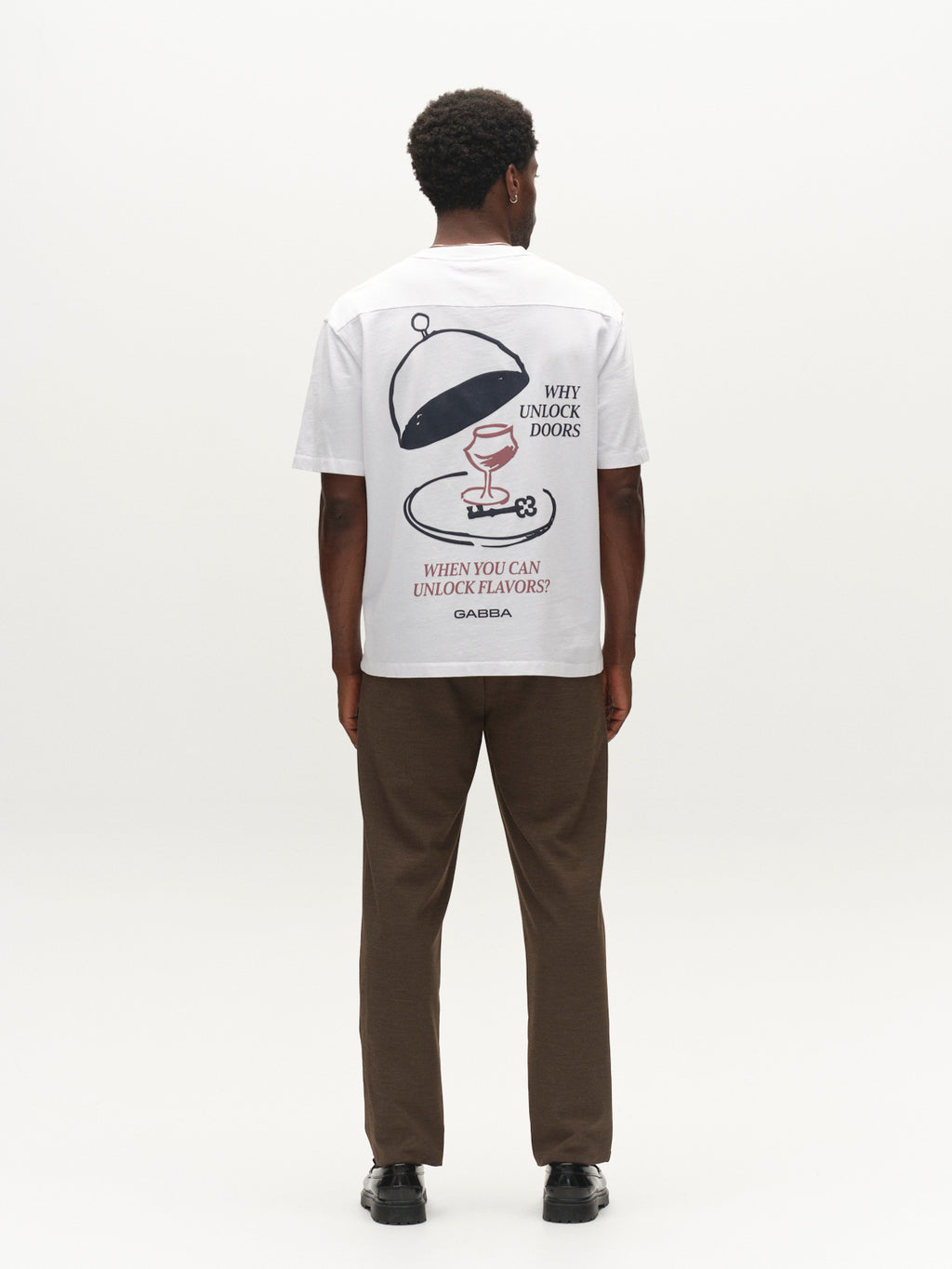 A man stands facing away, wearing a white T-shirt with a graphic of a cloche, a key, and the text “Why unlock doors when you can unlock flavors? GABBA.” He pairs it with Gabba’s GABnapoli 19 Phin GRS - Crocodile brown wool blend chinos in a regular straight fit and black shoes.