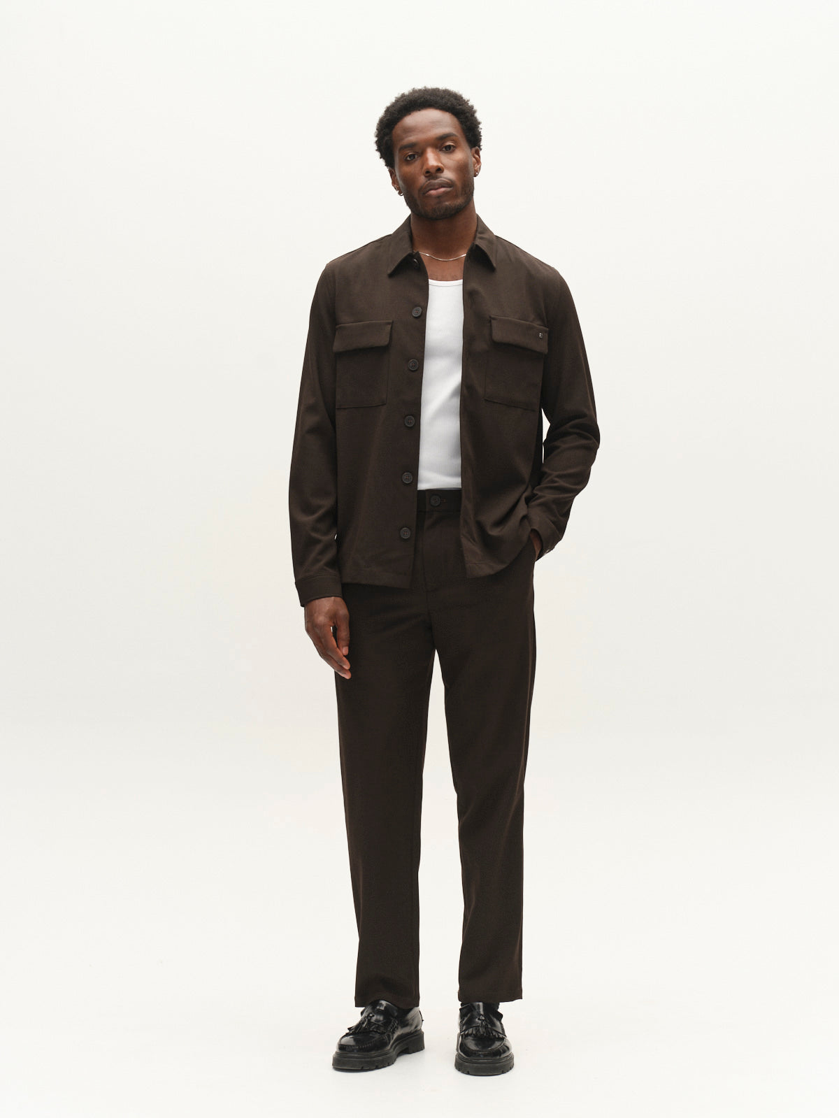 A man stands against a plain white background wearing Gabba’s GABnapoli 19 Phin GRS chinos in Brown Slate—a regular straight fit wool blend made with recycled polyester—paired with a dark brown jacket over a white shirt and black shoes.