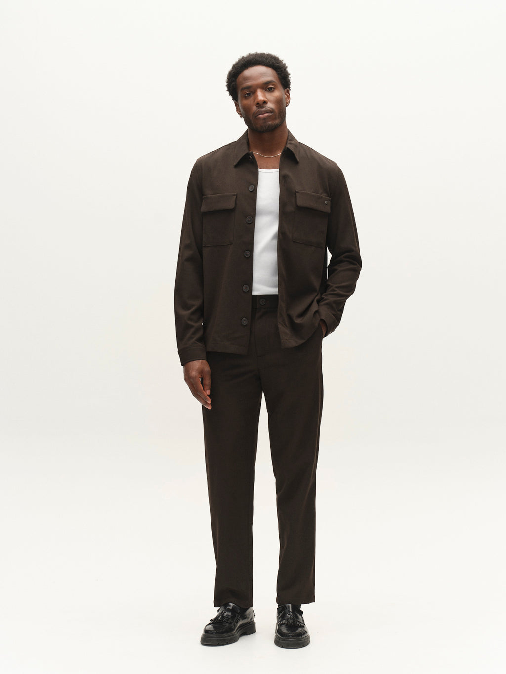 A man stands against a plain white background wearing Gabba’s GABnapoli 19 Phin GRS chinos in Brown Slate—a regular straight fit wool blend made with recycled polyester—paired with a dark brown jacket over a white shirt and black shoes.