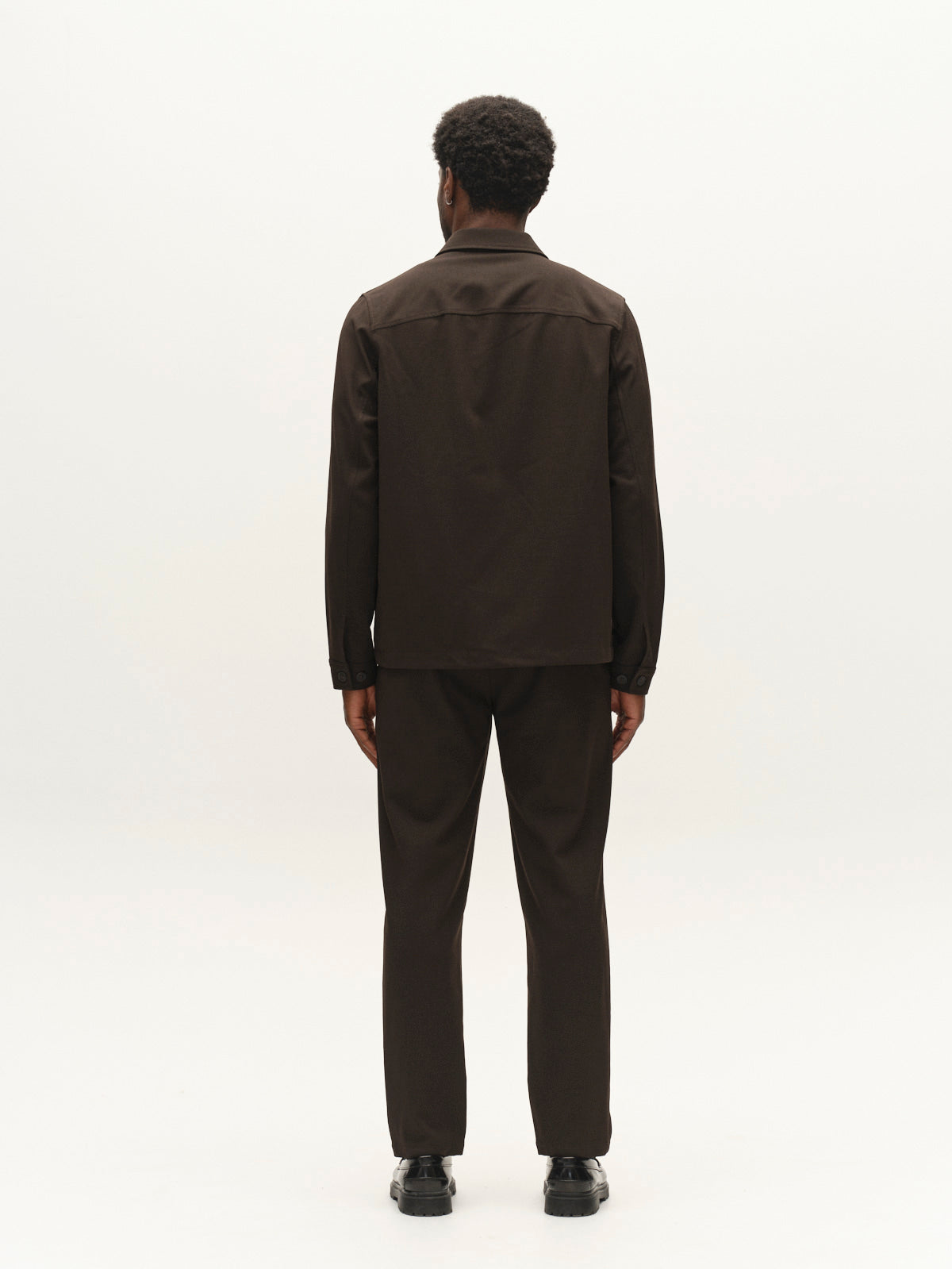 A man with short curly hair stands facing away, wearing the Gabba GABnapoli 19 Phin GRS - Brown Slate wool blend chinos and a dark jacket with black shoes, against a plain white background.