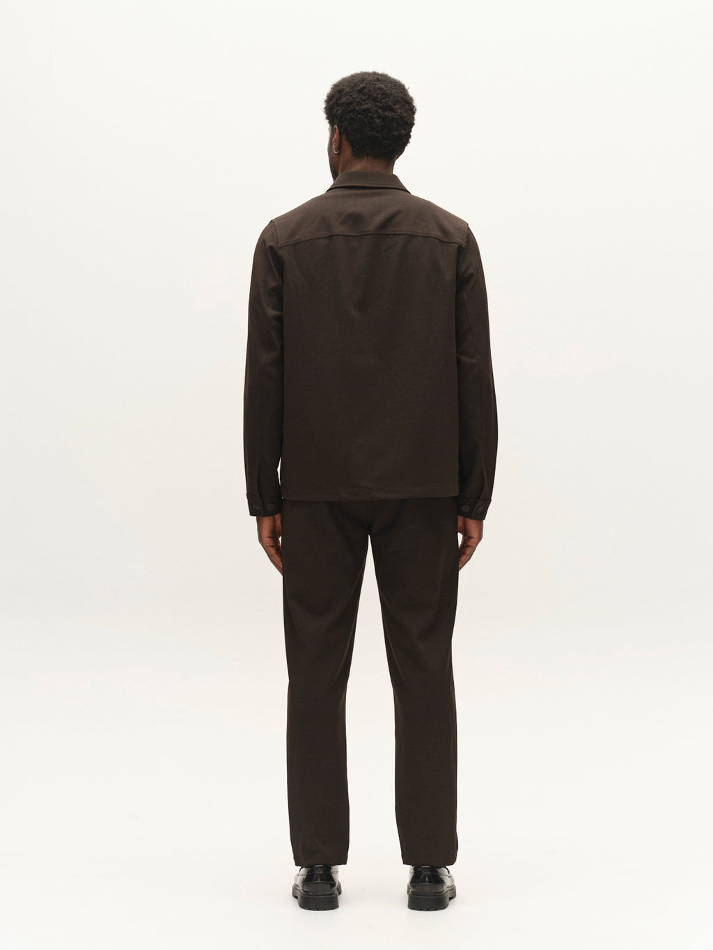 A man with short curly hair stands facing away, wearing the Gabba GABnapoli 19 Phin GRS - Brown Slate wool blend chinos and a dark jacket with black shoes, against a plain white background.