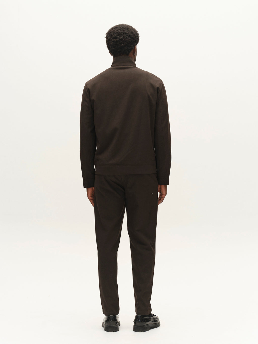 A person with short, curly hair stands facing away against a plain white background, wearing Gabba’s GABnik Phin GRS - Brown Slate: a men’s regular fit jacket and matching pants crafted from a wool and viscose blend, paired with black shoes—a subtle statement in modern outerwear.