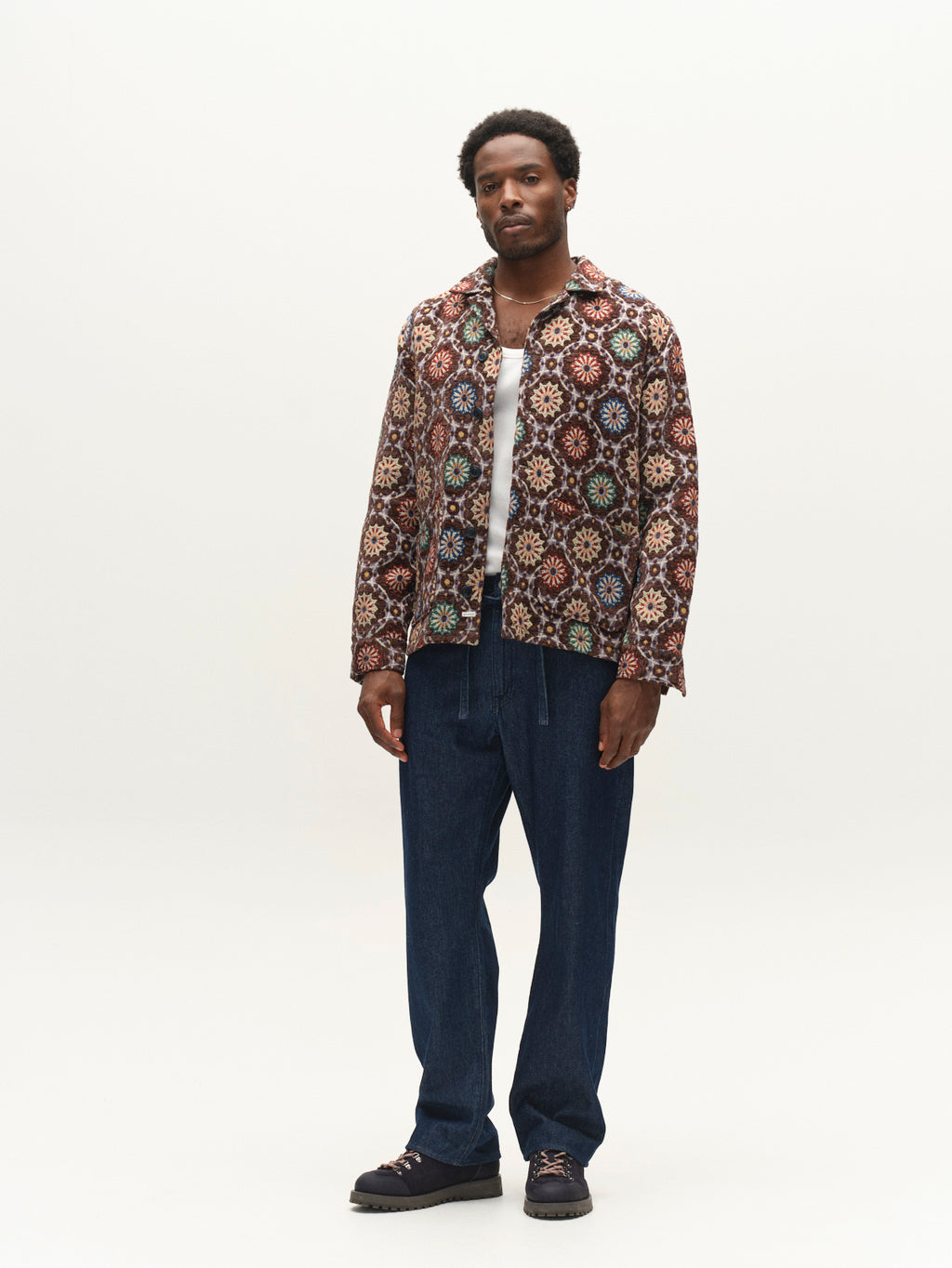 A man stands in a studio wearing the Gabba GABsimone Nava LS jacket in brown, crafted from textured jacquard fabric, paired with a white shirt, blue pants, and dark shoes. He looks at the camera against a plain light background.