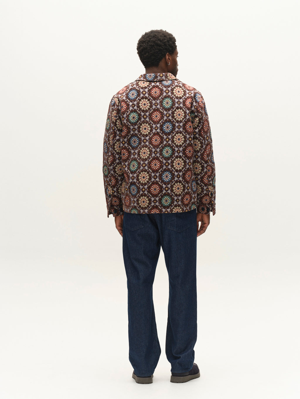 A person stands facing away, wearing the Gabba GABsimone Nava LS jacket in brown, featuring a colorful abstract motif and a resort collar, paired with loose-fitting blue jeans against a plain white background.