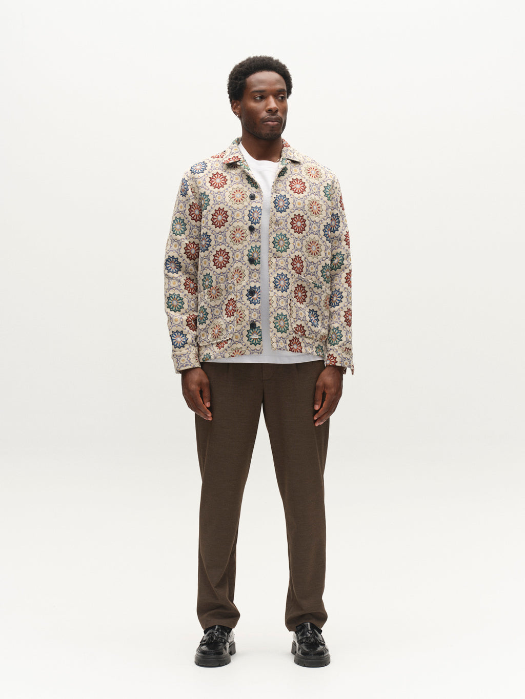 A man stands facing forward, wearing the Gabba GABsimone Nava LS - Birch jacket featuring a colorful floral jacquard pattern, paired with relaxed-fit brown pants and black shoes, against a plain white background.