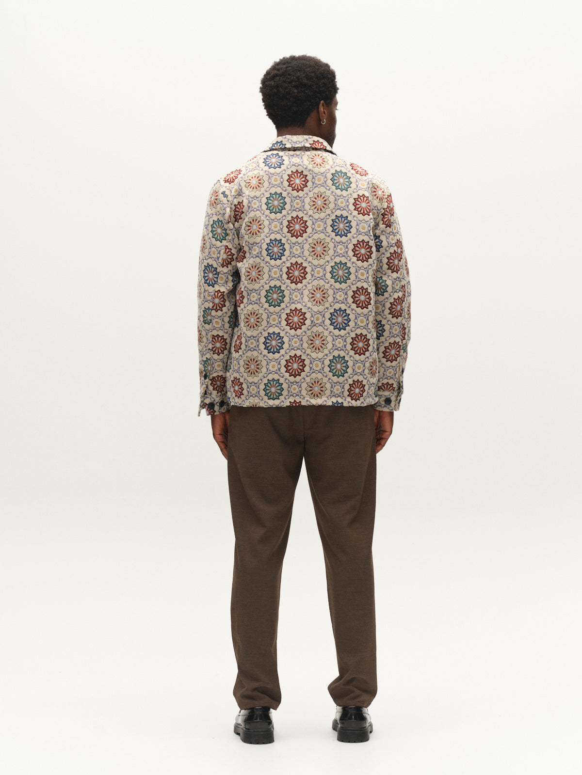 A person stands facing away, wearing the Gabba GABsimone Nava LS - Birch, a relaxed fit jacquard jacket featuring colorful circular patterns, paired with brown pants. The background is plain and white.
