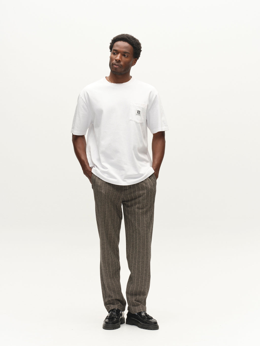 A man stands against a plain white background wearing a white t-shirt paired with Gabba’s Napoli 19 AX Stripes pants in dark grey, featuring brown pinstripes, a relaxed fit, elasticated waistband, and chino pocket construction. His hands are in his pockets as he looks slightly to the side with a neutral expression.