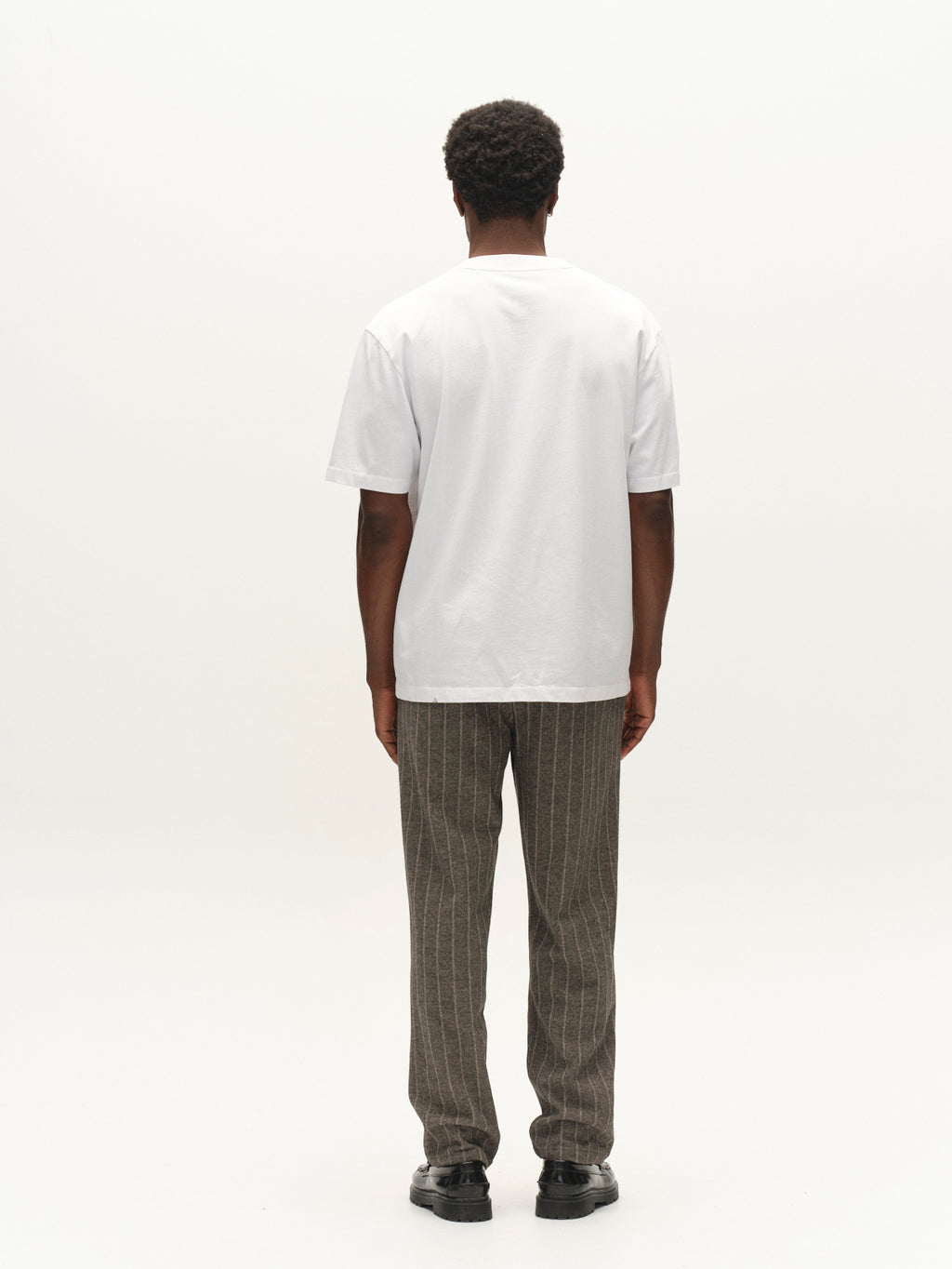 A person stands facing away from the camera, wearing a plain white T-shirt, Gabba Napoli 19 AX Stripes - Dark Grey relaxed fit pants featuring brown pinstripes with chino pocket construction and an elasticated waistband, and black shoes, against a plain white background.