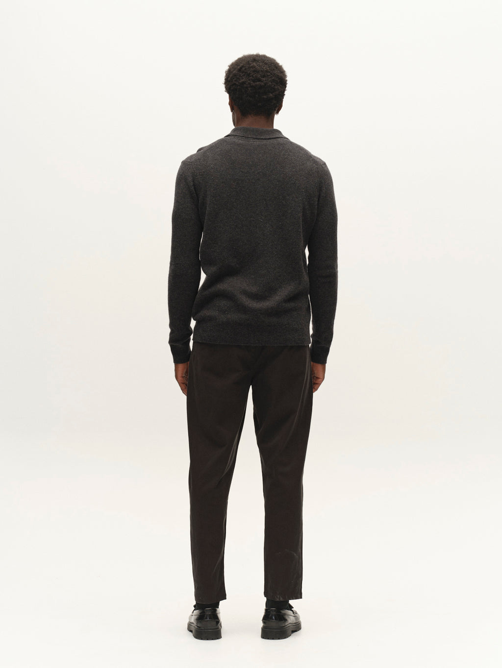 A man stands facing away from the camera, wearing a dark long-sleeve sweater, black shoes, and Gabba’s GABfirenze Bolu Cropped TENCEL™ Pant in Black, against a plain light background.