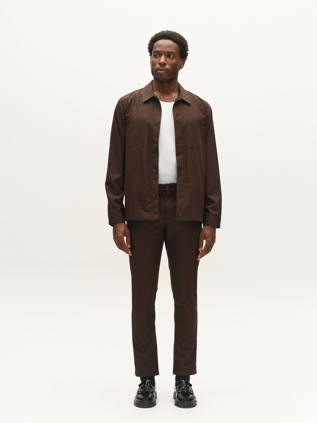 A man stands against a plain white background, wearing a white shirt under a dark brown button-up jacket, paired with Gabba’s GABpaul Deme Pant in Brown Slate and black shoes.