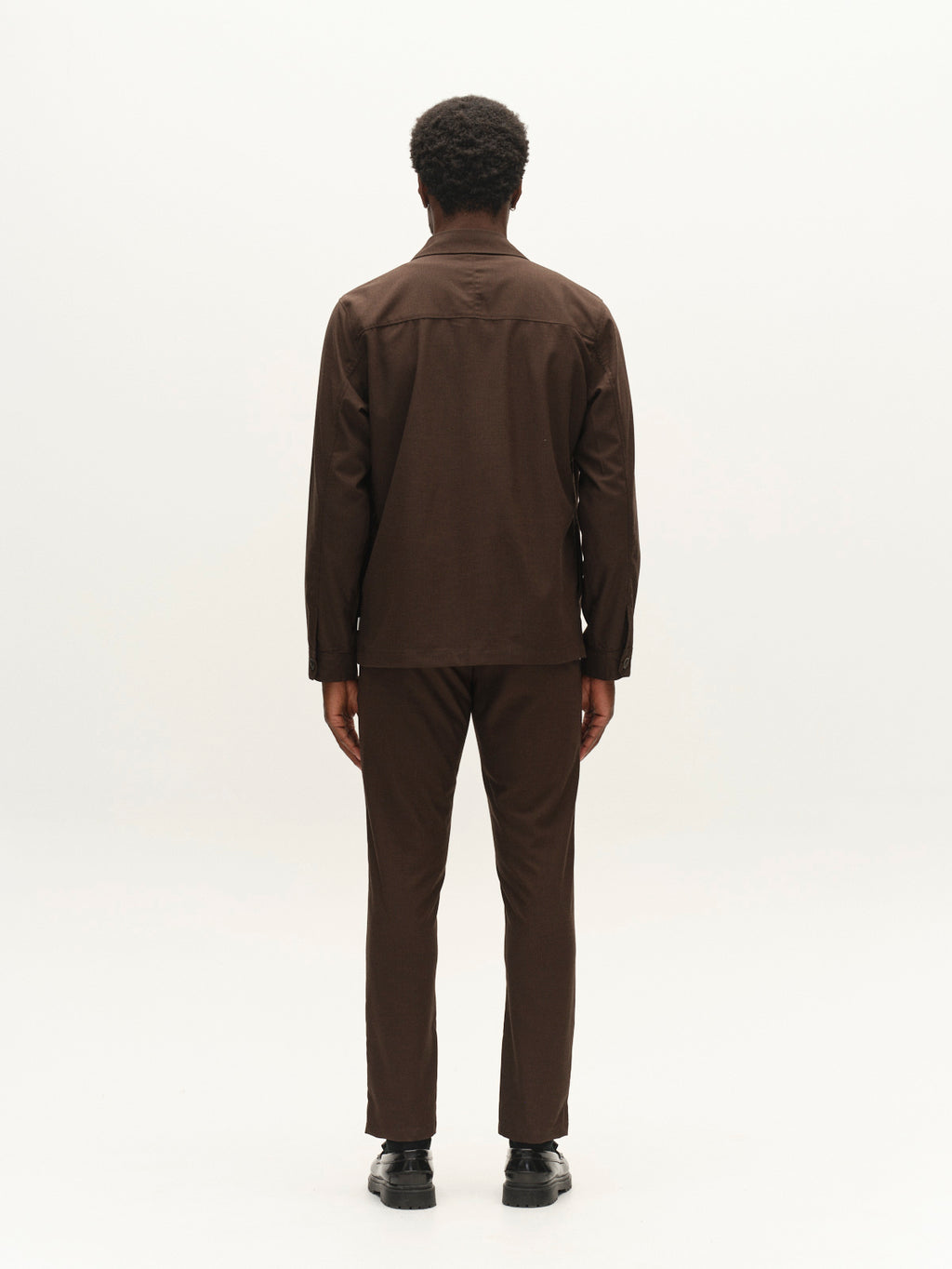 A person with short curly hair stands facing away, wearing a matching dark brown long-sleeve shirt and Gabba’s GABpaul Deme Pant in Brown Slate, styled in a slim chino fit and paired with black shoes, against a plain white background.