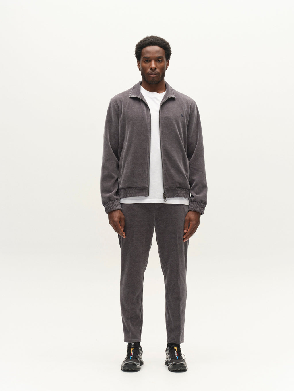 A man stands facing forward, wearing the Gabba GABnik Pulz Fune Jacket in Turbulence Blue with a full zip closure, matching gray pants, a white t-shirt, and black sneakers, against a plain white background.